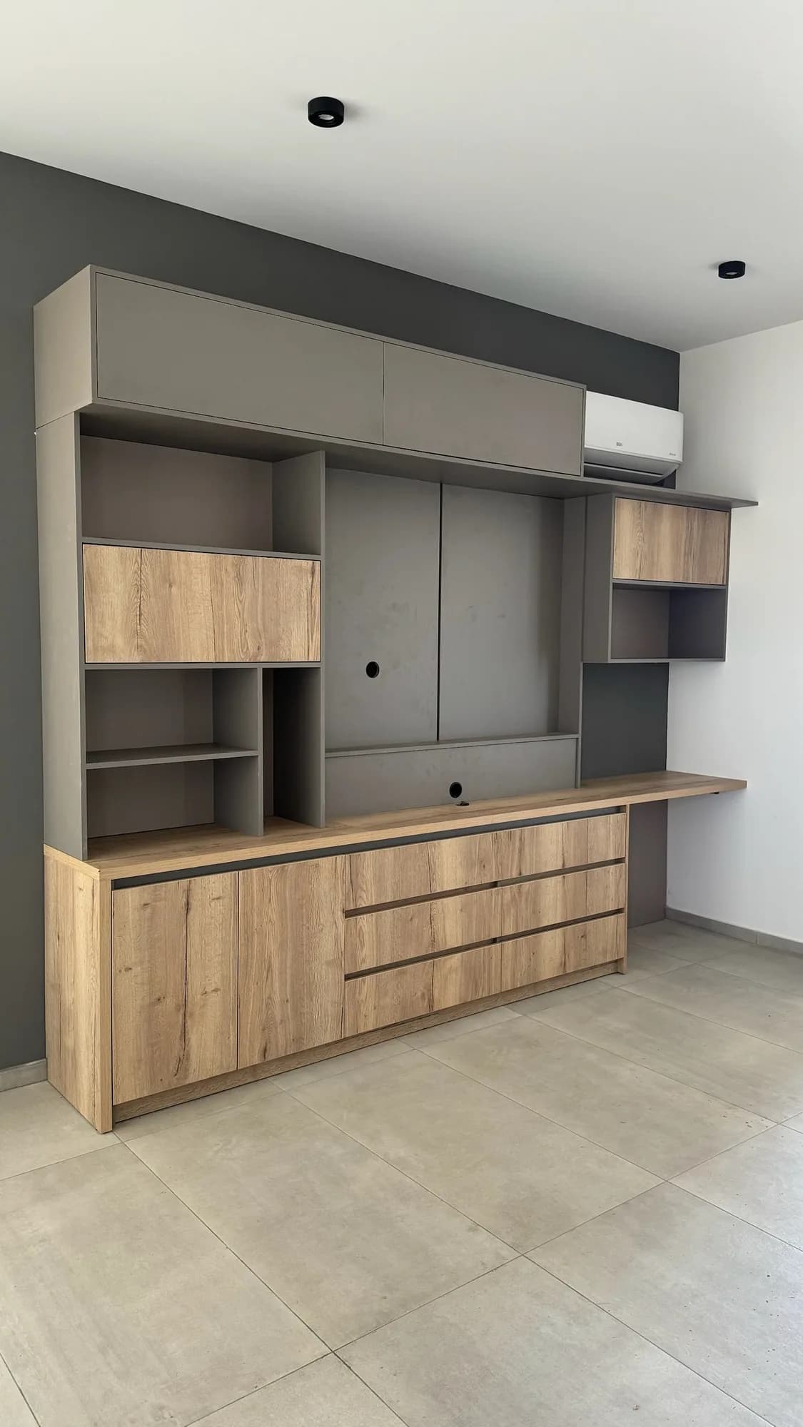 Mueble de living en melamina roble y gris topo con panel para TV, alacenas altas, nichos, cajonera con ranuras y escritorio volado; toma en diagonal desde la izquierda
