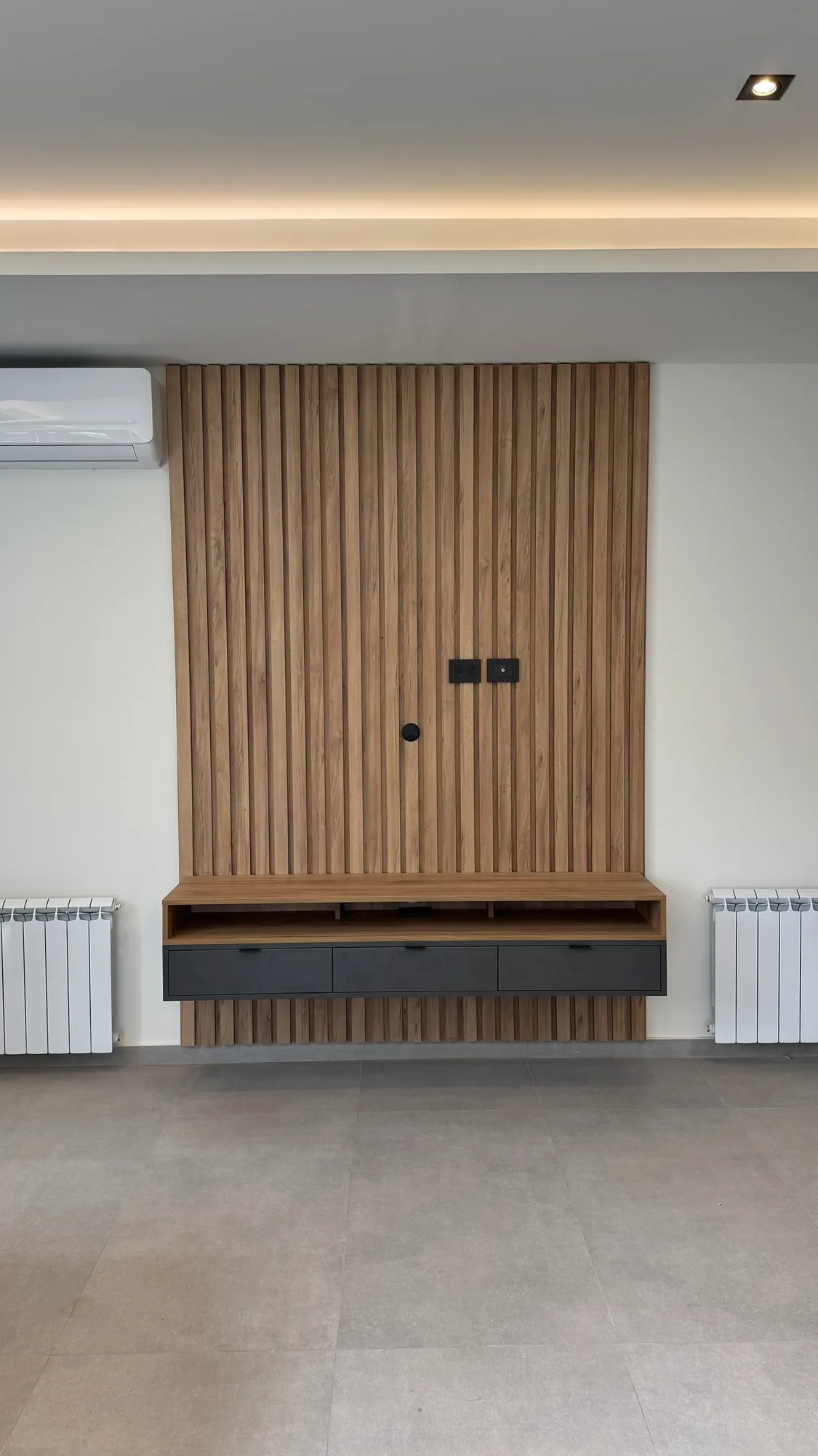 Panel de TV con listones de madera y rack flotante de tres cajones gris oscuro sobre pared clara, entre dos radiadores; toma frontal vertical con cielorraso con luz LED indirecta