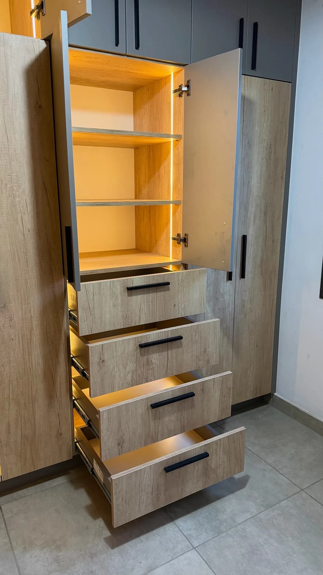 Columna de placar con puerta abierta, estantes con tira LED y cajonera de cuatro cajones extraíbles abiertos con tiradores negros; toma vertical sobre piso porcelanato
