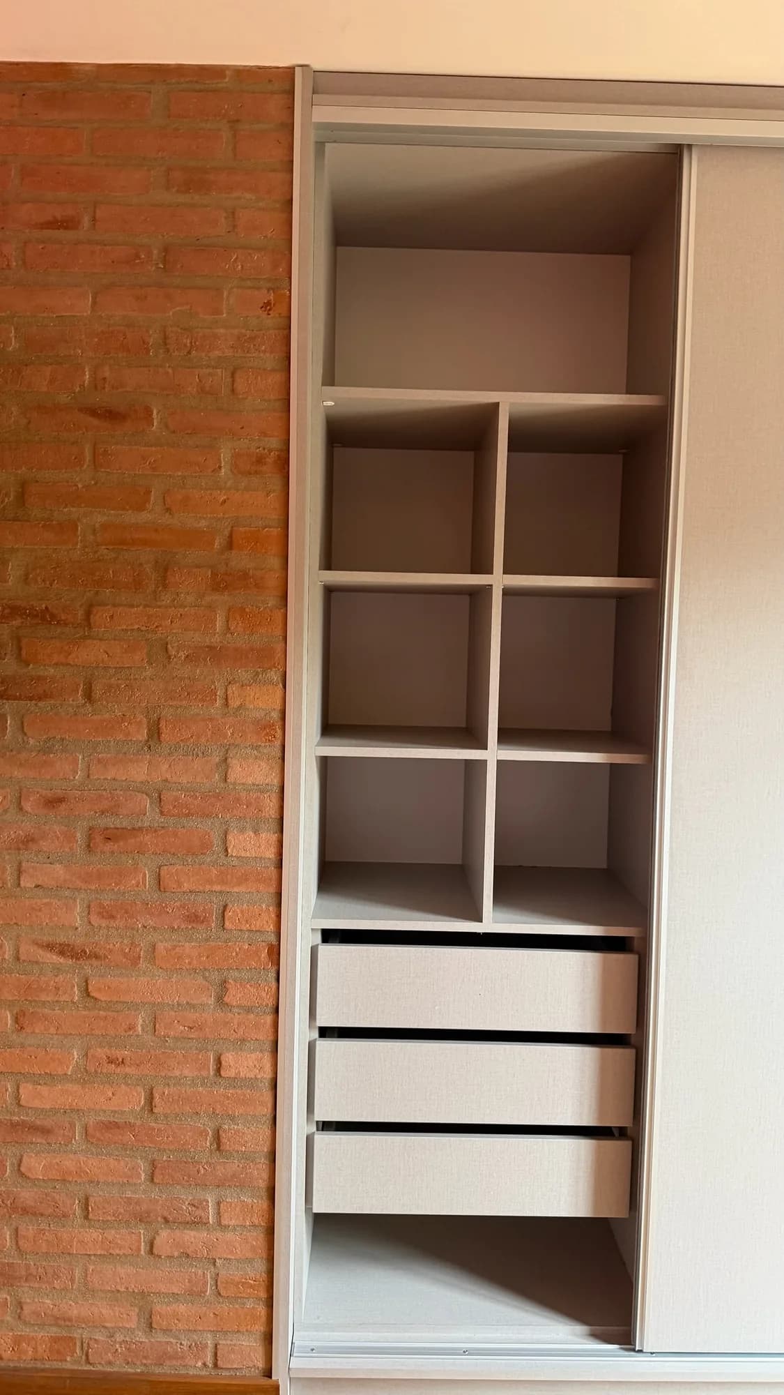 Placard de melamina gris con seis nichos abiertos, tres cajones y puerta corredera, junto a una pared de ladrillo visto; toma frontal