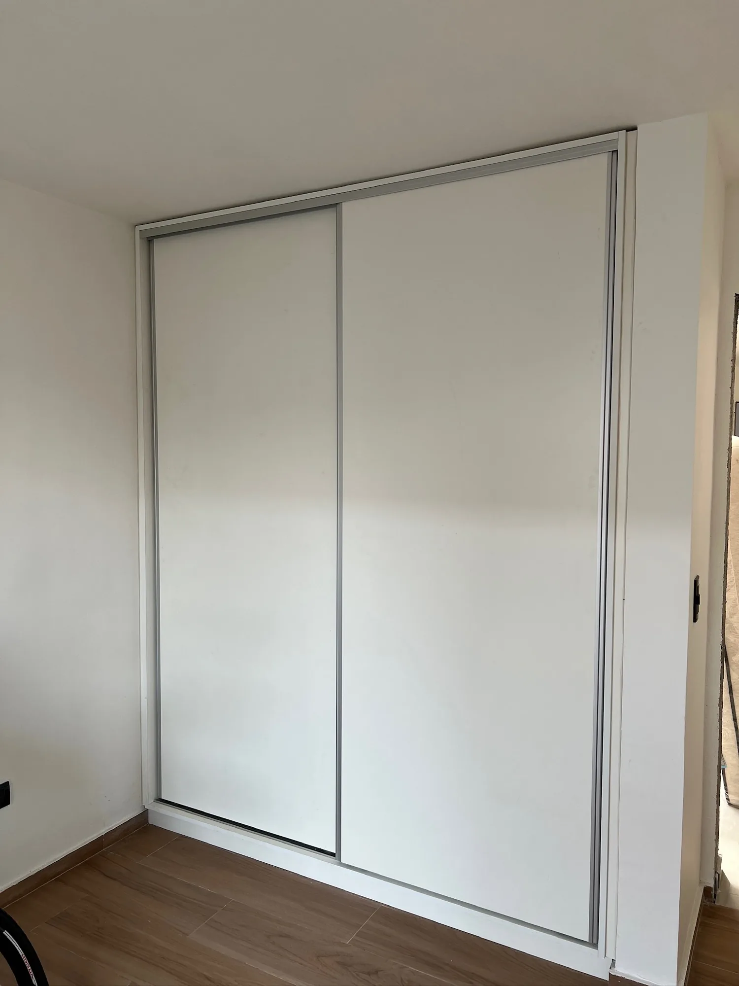 Placar empotrado de dos puertas corredizas blancas con perfilería de aluminio, en un dormitorio de piso símil madera; toma frontal
