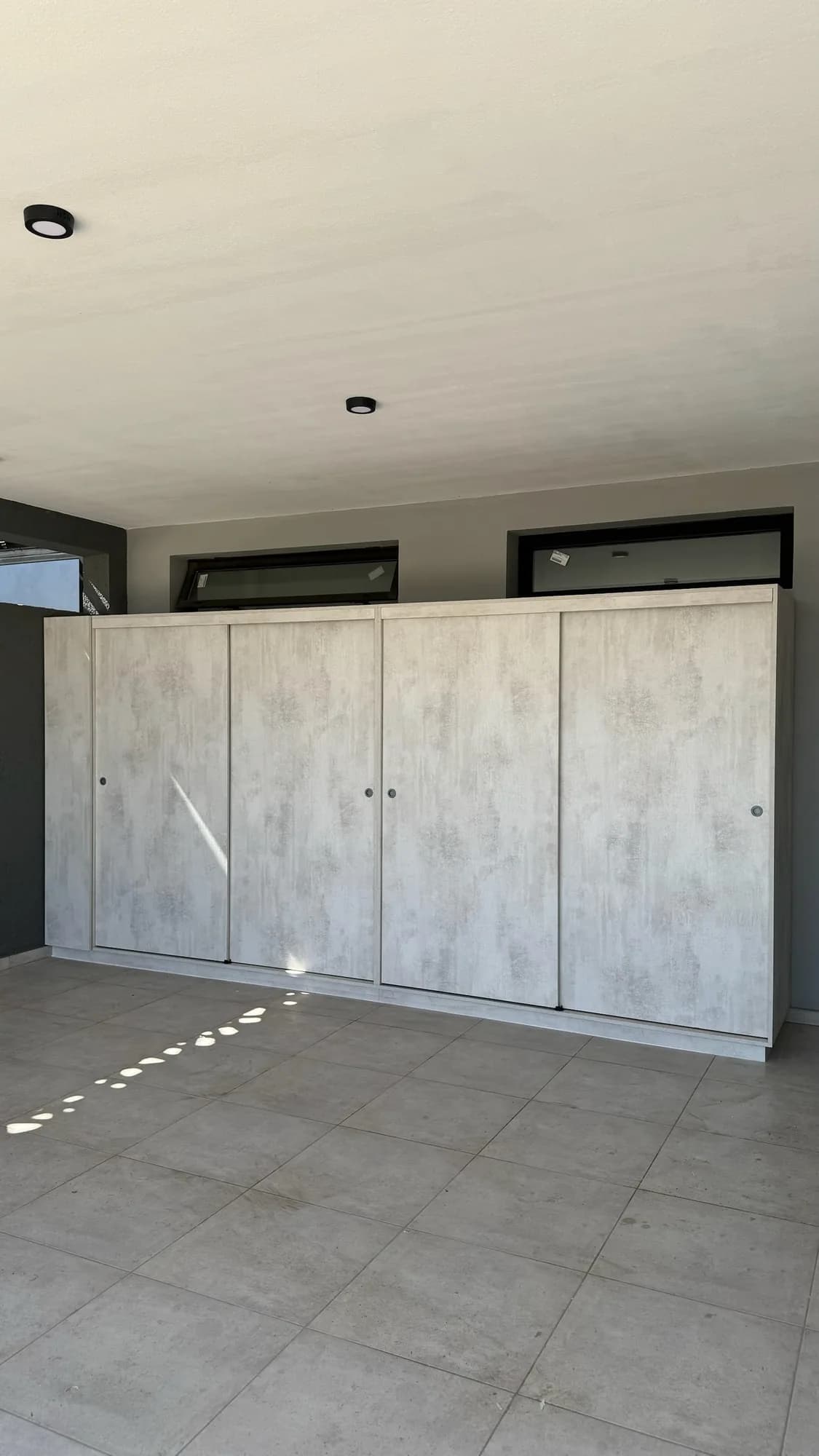 Mueble de cuatro puertas corredizas en melamina símil cemento claro con nichos abiertos en un extremo, apoyado contra una pared gris bajo ventanas altas; toma frontal en una galería cubierta