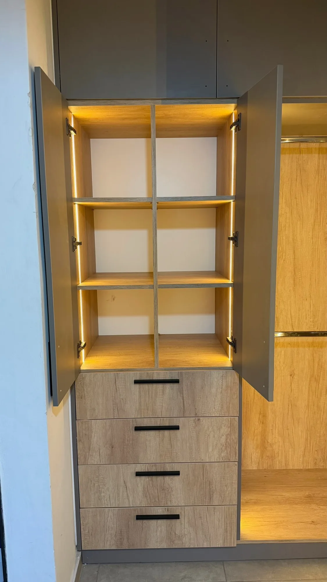 Módulo de placar con puertas grises abiertas, seis casilleros con tira LED y cajonera de cuatro cajones símil roble con tiradores negros, junto a un nicho de vestidor con barral; toma vertical frontal