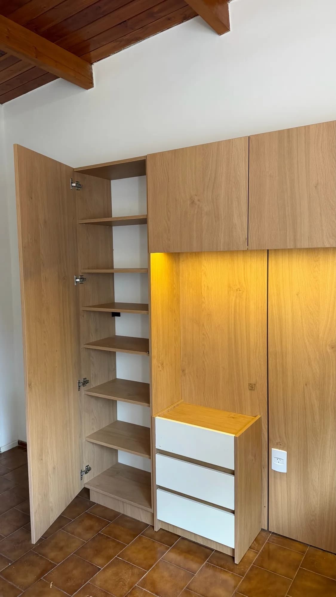 Placard en melamina símil roble con una puerta abierta que muestra estantes, junto a una cajonera blanca y un nicho con luz LED; toma lateral