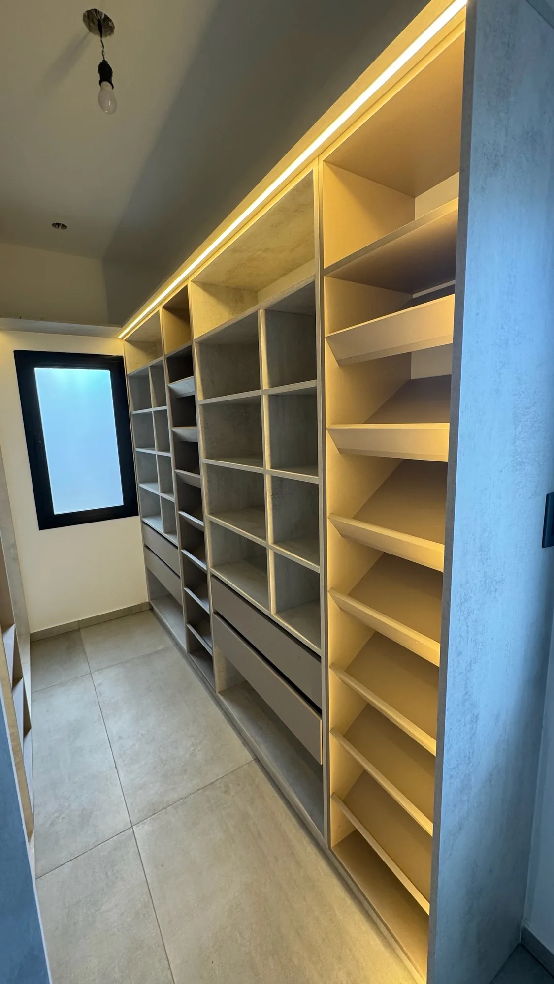 Vestidor pasillo con módulos de cubos, cajones y estantes inclinados para calzado en melamina símil cemento, con tira LED cálida en el frente superior y ventana al fondo; toma en perspectiva