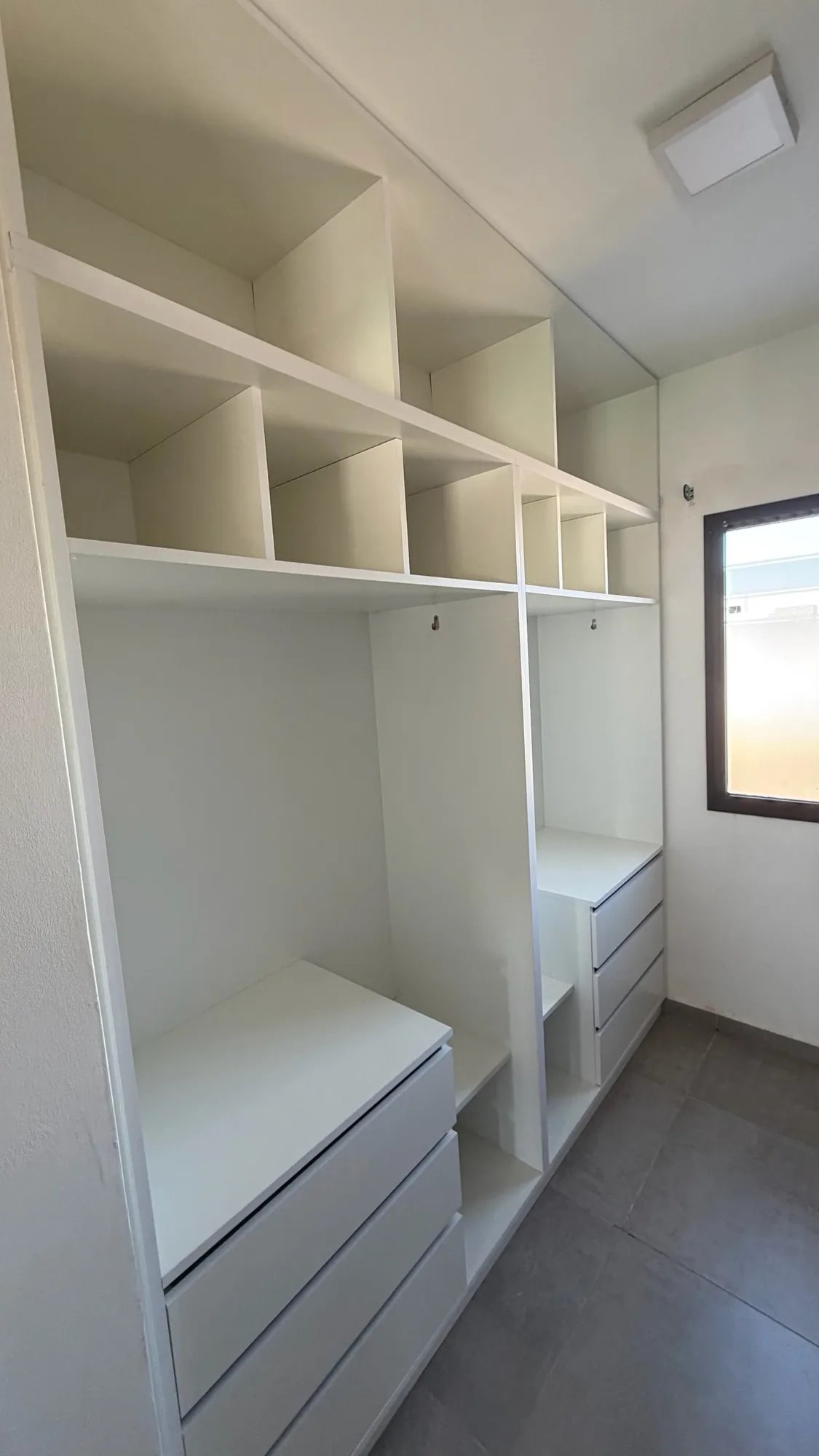 Vestidor tipo pasillo en melamina blanca hasta el techo, con estantes altos, divisiones, zona de colgado y dos cajoneras de tres cajones. Entra luz por una ventana al fondo; toma en gran angular