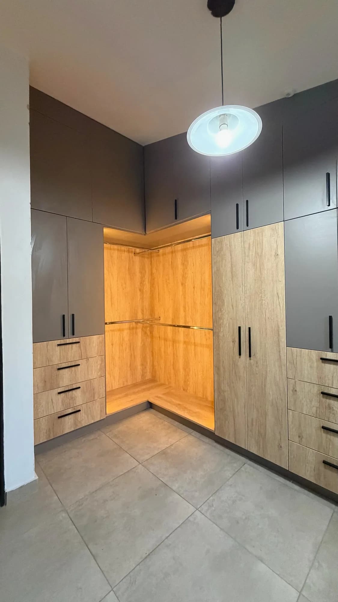 Vestidor en esquina con placares en gris y símil roble, cajoneras con tiradores negros y nicho abierto con barrales y luz LED cálida; toma vertical hacia la esquina