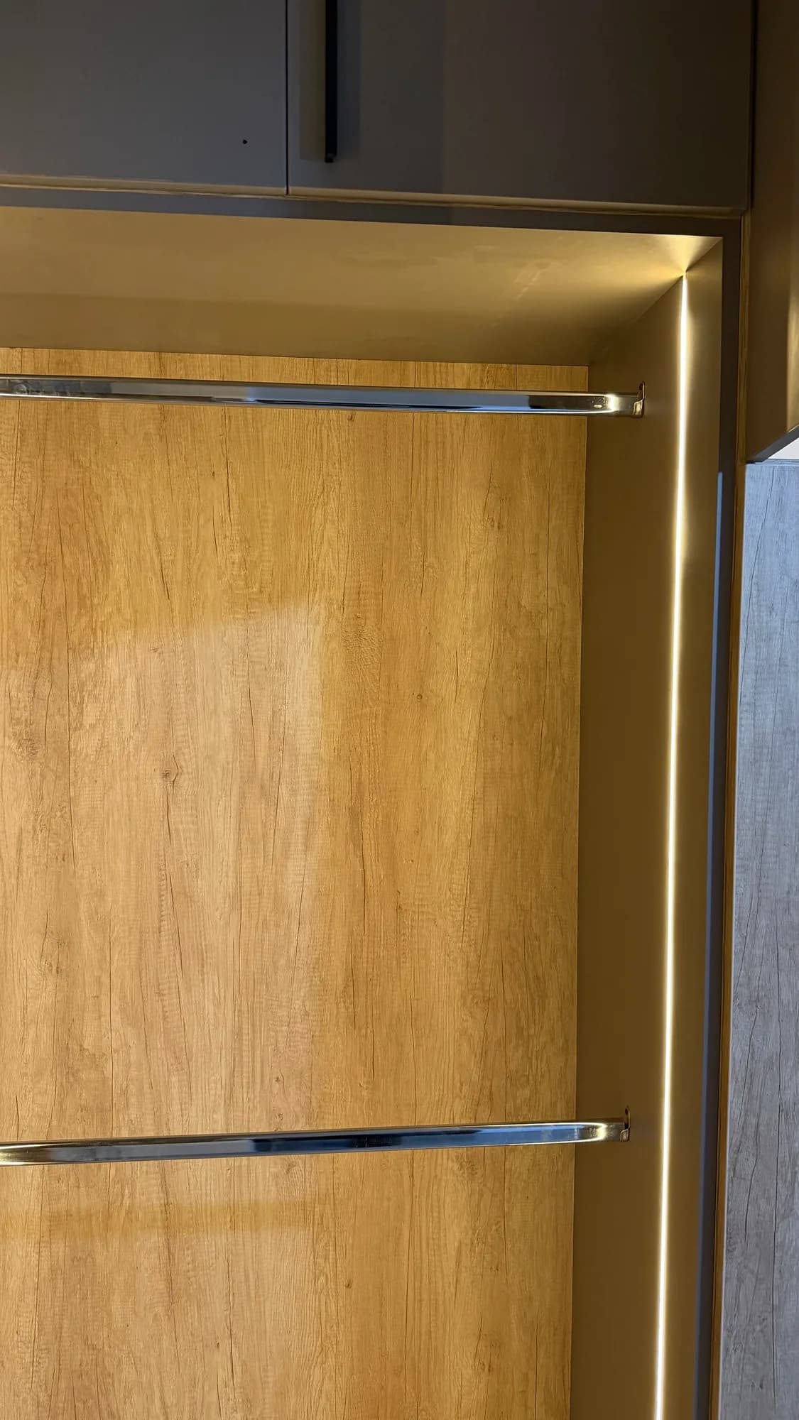 Nicho de vestidor con fondo símil roble, dos barrales cromados y tira LED lateral, bajo alacenas grises; toma vertical cercana