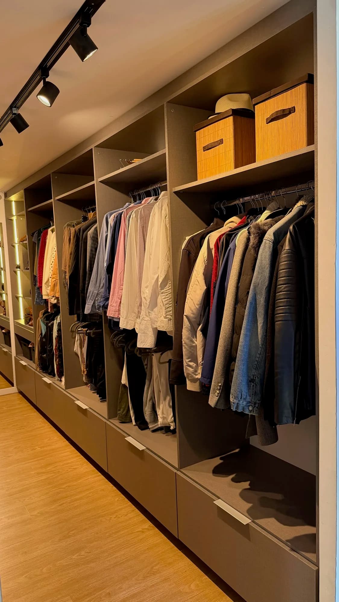 Vestidor con módulos abiertos en melamina gris, barrales con camisas y camperas, cajas de bambú en el estante superior y cajones con tiradores de aluminio; toma cercana en diagonal con spots sobre riel