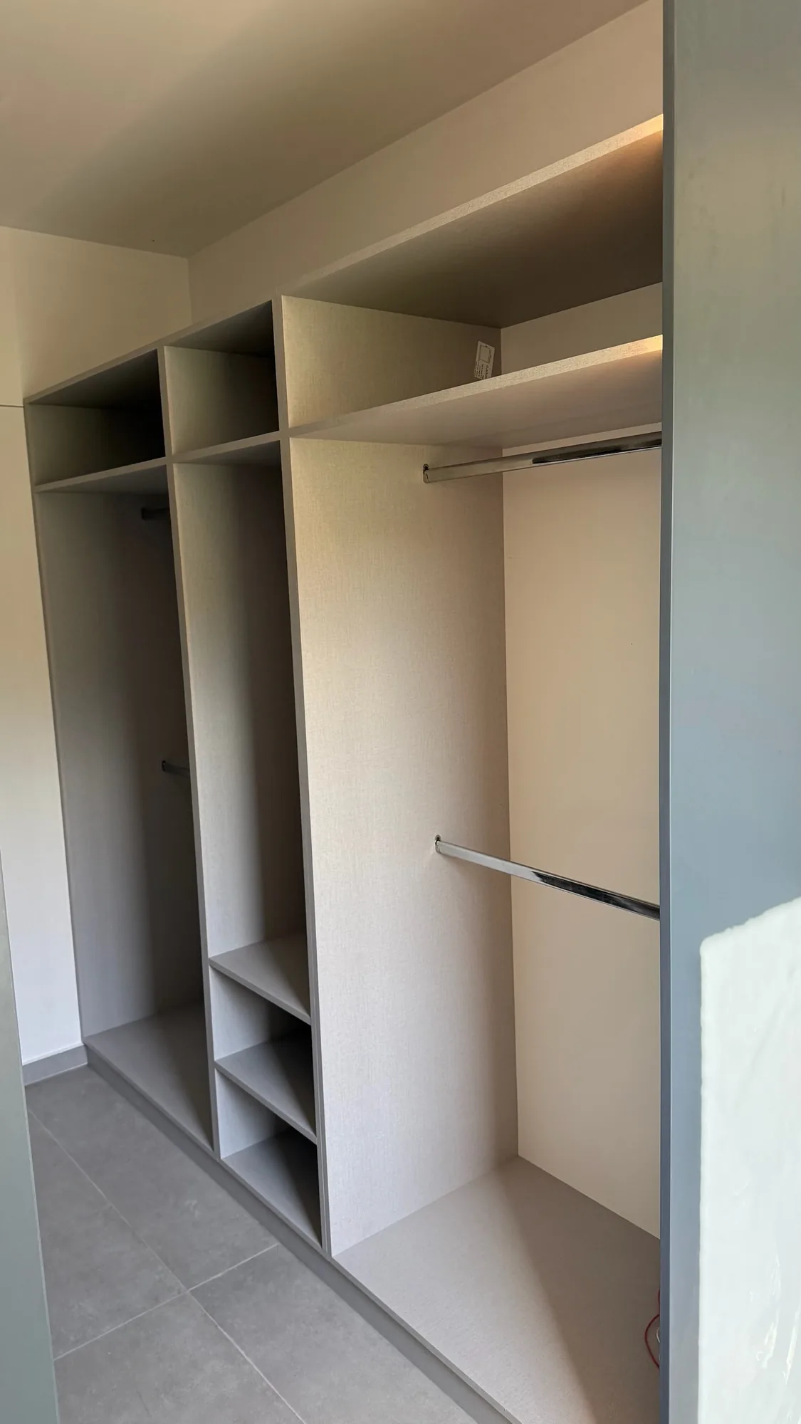 Vestidor abierto con barrales cromados, estantes y cajoneras en melamina gris beige con textura de tela, luz LED indirecta bajo los estantes altos y lateral gris antracita; toma en diagonal